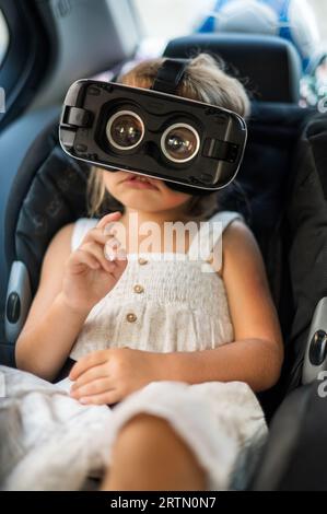 Kleines Mädchen, das im Auto in VR-Brille sitzt, ohne Vorderabdeckung im Kindersitz Stockfoto