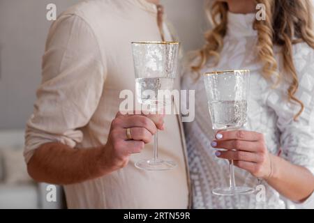 Das Paar hält und toastet Champagnergläser. Nahaufnahme der Hände, die trendige Gläser mit Wein halten. Einfarbige beige Kleidung. Gemütlicher Stil. Liebhaber machen es Stockfoto