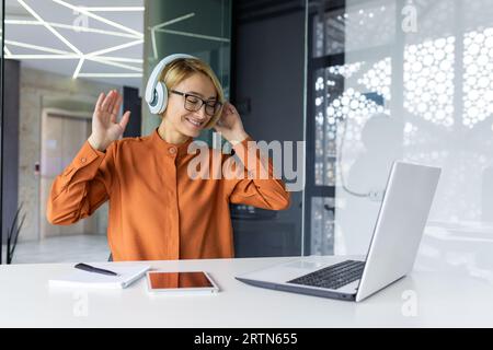 Junge, freudige Frau am Arbeitsplatz hört Musik in Kopfhörern, Geschäftsfrau tanzt und singt, sitzt am Arbeitstisch im Büro, Programmiererin gut gemacht erfolgreiche technische Projekt feiert. Stockfoto