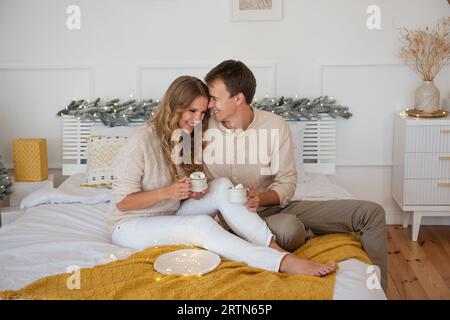 Niedliches, fröhliches Paar, das sich an Weihnachten mit gelber Decke auf dem Bett kuschelt. Zarte Frau und junger Mann trinken Kakao mit Marshmallows aus weißen Tassen. Mühle Stockfoto