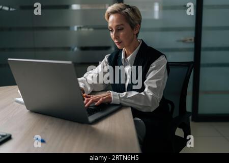 Mittlere Aufnahme einer fokussierten Geschäftsfrau mittleren Alters, einer CEO-Mitarbeiterin im Anzug, die am Schreibtisch sitzt und auf einem Laptop schreibt und auf den Bildschirm des Geräts schaut Stockfoto