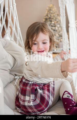 Nahaufnahme eines lustigen kleinen Mädchens, das zu Hause auf einem Makramee-Stuhl auf einer Schaukel reitet. Kinderschaukel, die herumtanzt. Gemütliche Atmosphäre. Rustikaler Stil Stockfoto