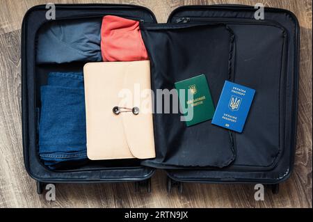 Reisetasche auf Rädern mit farbiger Kleidung, Ordner und ukrainischem Pass öffnen. Grüne Buchübersetzung: "Militärische ID". Grenze, Tourismus Stockfoto