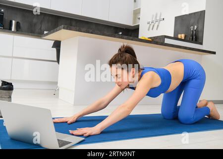 Porträt einer Frau, die Dehnübungen macht, folgt dem Tutorial auf dem Laptop, nimmt an Online-Yogastunden von zu Hause aus Teil, trainiert auf einer Gummimatte Stockfoto