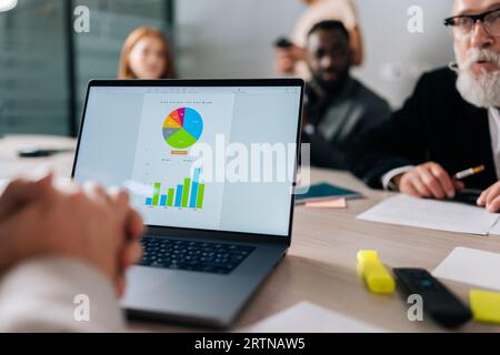Nahaufnahme vom Laptop mit Diagramm bis hin zur Gruppe verschiedener Geschäftsleute, die sich im Sitzungssaal des Büros treffen, um Geschäfte zu lösen und die Zusammenarbeit mit Partnern zu planen Stockfoto