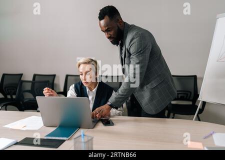 Erfahrene, mittelalte, kaukasische CEO-Managerin der 50er Jahre, die Anzug trägt und jungen, afroamerikanischen Praktikanten betreut. Stockfoto