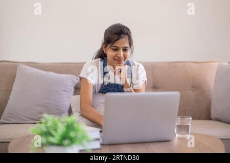 Junge indianerin lächelt mit Laptop zu Hause. Selbstbewusste ethnische Frau, die auf einem Wohnzimmertisch mit Laptop sitzt Stockfoto