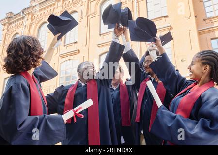 Eine multiethnische Gruppe von Studenten in Gewändern, die ihren Hut aufrichten und ihren Abschluss feiern Stockfoto