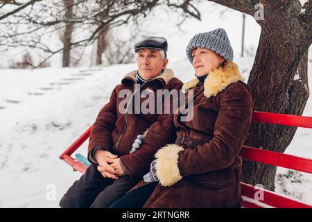 Seniorenpaar sitzt bei schneebedecktem Winterwetter draußen auf einer Bank. Ältere Rentner, die im Park chillen, bewundern die Aussicht in warmer Kleidung Stockfoto