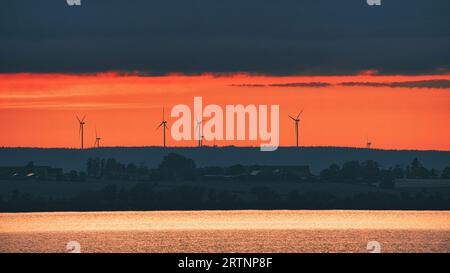 Windkraftanlagen, erneuerbare Energien, auf einem See bei Sonnenuntergang. Sauberer Strom für den Klimawandel. Bewölkter Himmel mit orangenem Sonnenschein in Skandinavien. Stockfoto