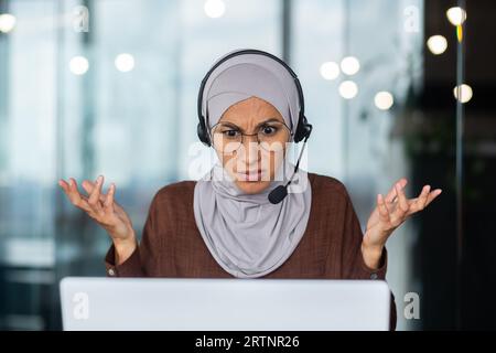 Nahaufnahme. Ernsthafte, strenge und anspruchsvolle muslimische Lehrerin in Hijab und Headset, die online über Laptop erklärt und unterrichtet. Stockfoto
