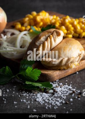 Eine Auswahl an frisch zubereiteten Empanadas auf einem Holzbrett, serviert mit gehackten Zwiebeln und Mais Stockfoto