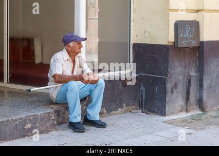 Havanna, Kuba - 30. August 2023: Ein kubanischer Mann mit einem Stock, der vor der Haustür eines Unternehmens sitzt Stockfoto