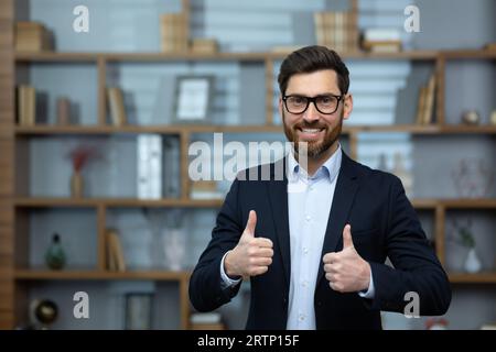 Porträt eines erfolgreichen Finanziers im Büro, der Mann lächelt und schaut auf die Kamera, zeigt einen positiven Daumen nach oben, Stockfoto
