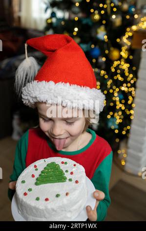 Kind in einem roten Hut mit einem Neujahrskuchen, der seine Zunge ausstreckt und versucht, die Creme zu lecken. Winterferien, weihnachtskonzept, viel Spaß, viel Spaß Stockfoto