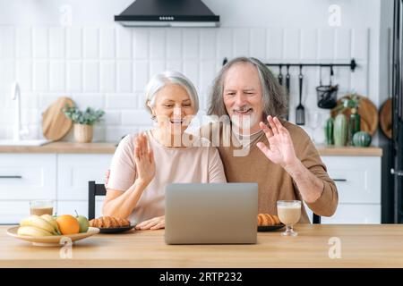 Glückliches weißes Rentnerpaar, Ehemann und Frau, sitzen zu Hause in der Küche, benutzen einen Laptop um über Online-Videokonferenz mit Kindern und Enkelkindern zu kommunizieren, begrüßen, fröhlich lächeln Stockfoto