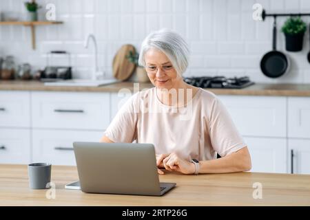Positive, moderne, attraktive, weiße, grauhaarige Frau, sitzt zu Hause in der Küche, verwendet ein Notebook für Fernarbeit, Online-Shopping, liest Nachrichten, sieht zufrieden aus, lächelt Stockfoto