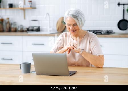 Glückliche weiße Dame mittleren Alters mit Brille, sitzt an einem Tisch mit einem Laptop zu Hause in der Küche, arbeitet online, sieht sich Videos an, liest Nachrichten, schaut auf den Laptop-Bildschirm, lächelt Stockfoto