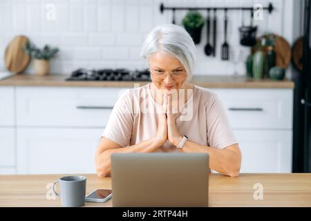 Hübsche, moderne, positive weiße, grauhaarige Frau mit Brille, die zu Hause in der Küche am Tisch sitzt, auf den Laptop-Bildschirm schaut, etwas will, sich Sorgen macht, Handflächen zusammenhält, betet Stockfoto