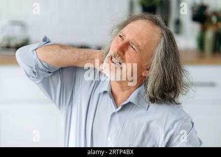 Nahaufnahme Foto eines ungesunden weißen älteren grauhaarigen Mannes, sitzt in der Küche am Tisch, fühlt sich überarbeitet, müde, hat Schmerzen im Nacken, massiert seinen Hals mit seinen Händen, Augen geschlossen, grimmig Stockfoto