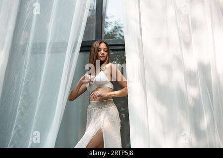 Anmutige und tätowierte Frau in Bikinioberteil und Rock in der Nähe von weißem Tüll aus Strandhaus Stockfoto