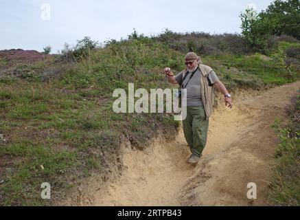 Stout-Mann von 70 mit einer Rohrwanderung auf einem Sandweg. Der Weg geht bergab und wird vom Regen ausgewaschen. Um ihn herum blüht das Heidekraut Stockfoto