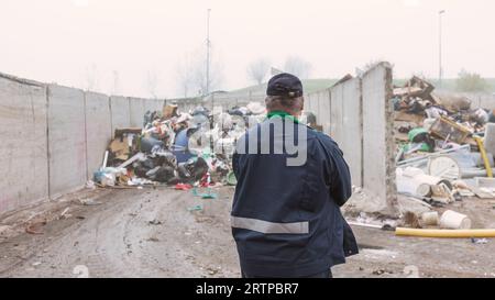 Ein Arbeiter im Recycling-Center, in dunkelblauer Arbeitskleidung, der sich einen unsortierten Müllhaufen ansieht, ist gerade in der Materialrückgewinnungsanlage angekommen, Rückansicht. Stockfoto