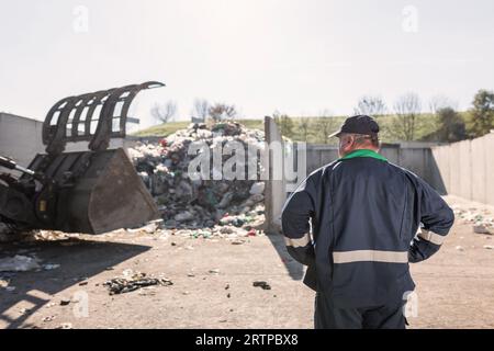 Ein Mitarbeiter des Recyclingzentrums in dunkelblauer Arbeitskleidung, der sich einen unsortierten Müllhaufen ansieht, ist gerade in der Materialrückgewinnungsanlage eingetroffen Stockfoto