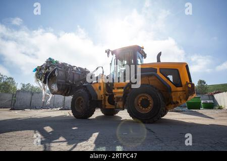 Gelber Radlader mit angehobenem Schrottgreifer, der sich im Bereich des Recyclingzentrums bewegt, während er entsorgten Abfall verarbeitet Stockfoto