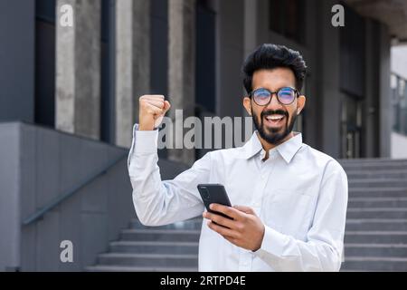 Arabisches Online-Gewinnerporträt, Mann im Business-Hemd, der durch die Stadt vor dem Bürogebäude läuft, Geschäftsmann, der lächelt und die Kamera anschaut, die Hand nach oben hält, die Triumphgeste verwendet die App am Telefon. Stockfoto