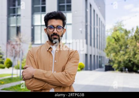 Ernsthaft denkender Geschäftsmann Programmierer, der Kamera mit gekreuzten Armen betrachtet, indischer Mann konzentrierte sich im Hemd vor dem Bürogebäude. Stockfoto