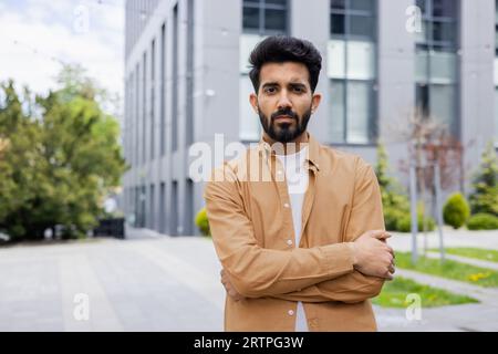 Ernsthaft denkender Geschäftsmann Programmierer, der Kamera mit gekreuzten Armen betrachtet, indischer Mann konzentrierte sich im Hemd vor dem Bürogebäude. Stockfoto
