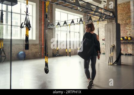Workout am Morgen. Rückansicht einer jungen Sportlerin, die ihre Sporttasche trägt und beim Stehen im leeren Fitnessstudio zur Seite schaut. Stockfoto