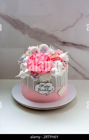 Wunderschöner und eleganter grau-rosa Kuchen mit geschmolzener weißer Schokolade, Makronen, essbarer Pfingstrosenblüte und Meringue-Küssen Stockfoto