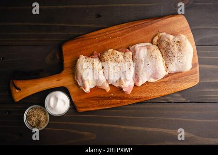 Gewürzte rohe Hähnchenschenkel mit Haut auf einem Holzschneidebrett: Ungekochte Stücke von dunklem Geflügel, gewürzt mit Meersalz und schwarzem Pfeffer Stockfoto
