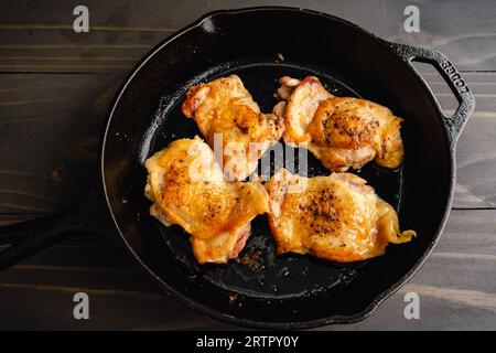 Gebratene Hähnchenschenkel in einer Eisenguss-Pfanne: Vier in der Pfanne gebratene Hähnchenschenkel mit Haut in einer Pfanne auf einem Holztisch Stockfoto