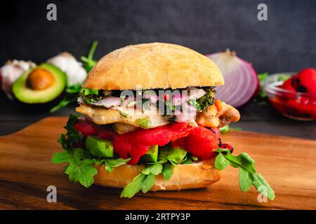 Chimichurri Hühnersandwich auf einem Holzbrett: Sandwich auf Ciabatta-Brot mit Avocado, gerösteten Paprika und Rucola Stockfoto