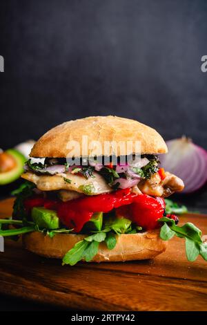 Chimichurri Hühnersandwich auf einem Holzbrett: Sandwich auf Ciabatta-Brot mit Avocado, gerösteten Paprika und Rucola Stockfoto