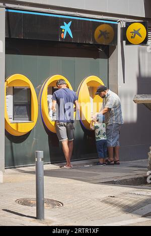 Viladecans, Spanien - 14. September 2023: Zwei Mann und ein Kind vor dem Geldautomaten Caixa ziehen an einem sonnigen Tag Geld ab. Stockfoto