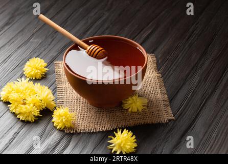 Honig in einer Tonschüssel, Dipper und gelbe Blumen auf Textil- und dunklem Holzhintergrund. Honigstäbchen liegen in einer Keramikschüssel mit Honignähe Stockfoto