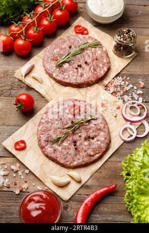 Rohe Burgerpasteten mit Gewürzen und Rosmarin auf Pergament, Sauce, frisches Gemüse, Hamburger-Zutaten auf Holzhintergrund. Stockfoto