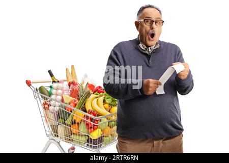 Schockierter reifer Mann mit einem Einkaufswagen, der eine Rechnung isoliert auf weißem Hintergrund hält Stockfoto