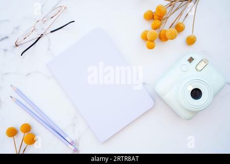 Flacher Tisch aus weißem Marmor mit Schreibwaren, Gläsern, gelben Blumen und Fotokamera. Draufsicht mit Kopierbereich. Modernes weibliches Arbeitsplatzkonzept Stockfoto