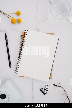 Flaches weißes Notizbuch mit Platz für Text, Gläser und gelbe Blumen auf weißem Marmorhintergrund. Draufsicht. Minimaler Schreibtisch im Home Office. Modern Stockfoto