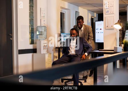 Sorglose Mitarbeiter, die Sportwettkämpfe im modernen Startup-Büro machen, Pause machen und Spaß haben. Fröhliche Angestellte in formellen Anzügen, die bis spät in die Nacht auf dem Schreibtisch sitzen Stockfoto