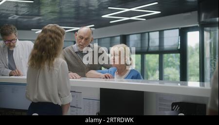 Ältere Ehepaare stehen in der Nähe der Rezeption im Lobby-Bereich der Klinik, kommen Sie nach einem Termin mit einem Arzt. Empfangsdame spricht mit Patienten am Informationsschalter. Die ärztliche Arbeit im modernen Krankenhaus. Stockfoto