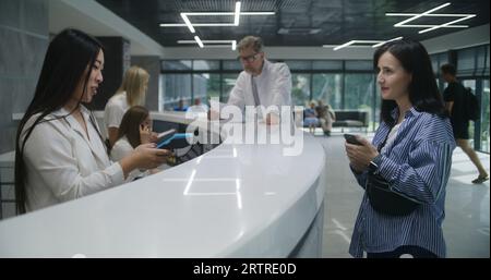 Asiatische Verwaltungsrätin an der Rezeption in der Klinik gibt dem Patienten das Terminal. Frau bezahlt kontaktlos für einen Arzttermin per Telefon. Medizinisches Personal arbeitet im medizinischen Zentrum. Stockfoto