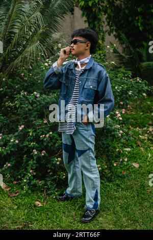 Ein asiatischer Mann mit Sonnenbrille und einem Jeanshemd, der mittags vor einer blühenden Pflanze steht Stockfoto