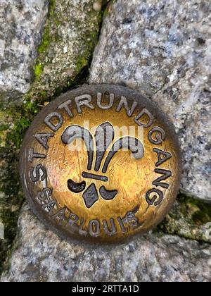 Messingfarbene Metallmedaille in einer kopfsteingepflasterten Straße. Die Medaille ist kreisförmig und hat ein Fleur-de-LIS-Symbol in der Mitte. Die Medaille ist es Stockfoto