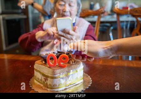 Ältere Frau macht Smartphone-Fotos von einem Kuchen, der ihren 90. Geburtstag feiert. Hände von Enkel zünden die Kerzen an. Stockfoto